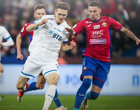 Футболисты в белой и красно-синей форме борются за мяч на поле. Один игрок пытается обойти другого, активная игра с напряжённой атмосферой. Вокруг видны другие участники матча и размытие толпы на заднем плане