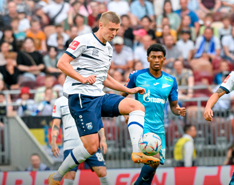 На изображении футболист в бело-синей форме контролирует мяч на поле, пока другой игрок в голубой форме наблюдает. На заднем плане видны зрители на стадионе