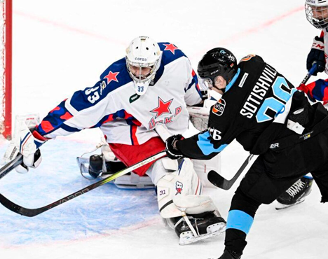 На изображении хоккейный вратарь в белой форме с эмблемой и красными звездами пытается отразить бросок игрока в черной форме с номером 36. Сцена происходит на льду перед воротами