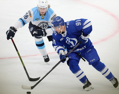 Хоккейный матч: два игрока в синей и белой форме на льду. Игрок в синем с шайбой, соперник в белом преследует. Оба в шлемах и коньках. Яркая динамика игры