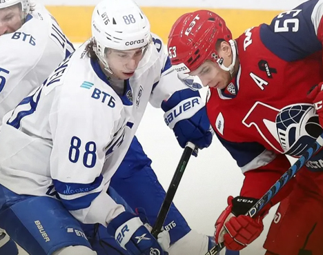 Хоккеисты в бело-синей и красной форме борются за шайбу на льду. Один игрок в белой форме, другой в красной, на шлеме у игрока в красной форме буква А. Спортивное состязание между двумя командами