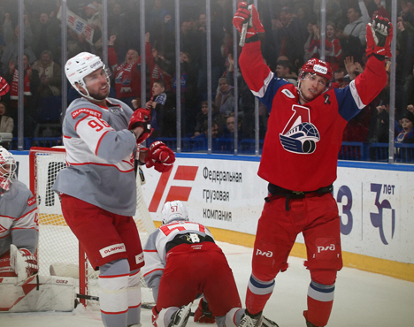 На изображении хоккеисты в красно-синей и серой формах на льду. Один игрок с поднятыми руками празднует гол, другие выражают разочарование. На заднем плане видны зрители и рекламные баннеры