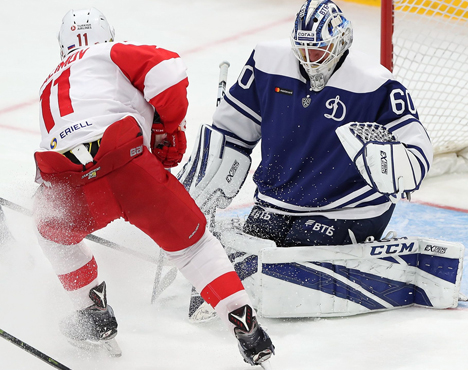 Хоккеист в красно-белой форме атакует ворота. Вратарь в синей форме пытается отразить шайбу, опустившись на колени. Оба игрока сосредоточены на игре, на льду виден снег от резкого движения