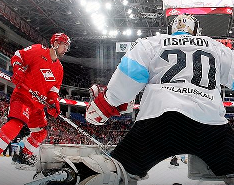 Хоккеист в красной форме атакует ворота. Вратарь в бело-голубой форме готовится отразить шайбу. На заднем плане трибуны с болельщиками и часть крыши арены