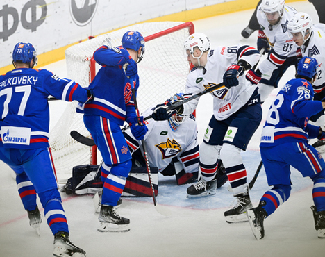 Хоккейный матч: игроки в форме синих и белых цветов борются у ворот. Вратарь в белой форме пытается предотвратить гол, игрок в синем делает попытку броска по шайбе. Интенсивная игра у ворот