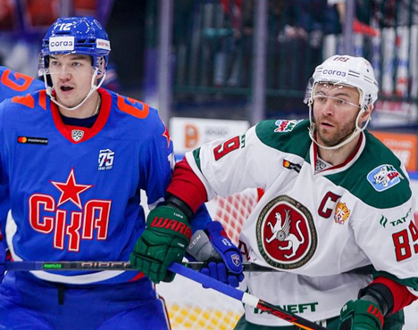 На изображении два хоккеиста на льду. Один в синей форме с красной звездой, другой в белой форме с эмблемой дракона. Они соревнуются за шайбу во время напряжённого матча