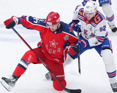 Хоккейное действие на льду: игрок в красной форме соревнуется с игроком в бело-синей форме. Они схлестнулись в напряженной борьбе за шайбу, оба в коньках и экипированы шлемами. На форме игроков видны логотипы и номера