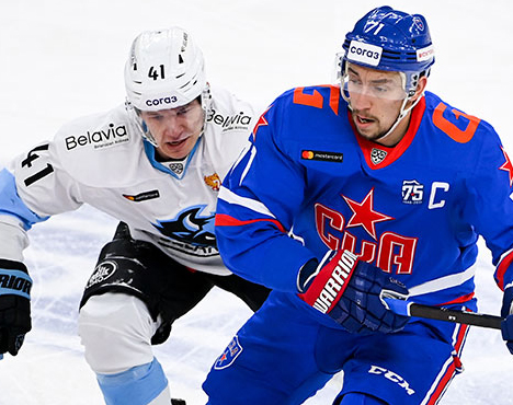 На изображении двое хоккеистов в динамике на льду: игрок в синей форме с эмблемой СКА и капитанской нашивкой и игрок в белой форме команды соперника. Эпизод матча в борьбе за шайбу