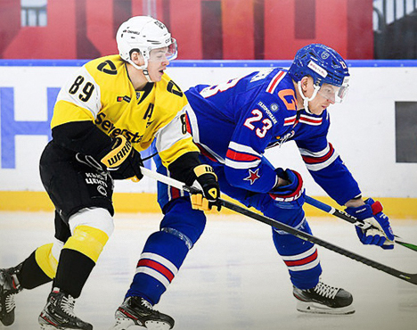 На изображении два хоккеиста в движении на льду. Один в желто-чёрной форме с номером 89, другой в синей с номером 23. Они борются за шайбу у борта с надписью на заднем плане