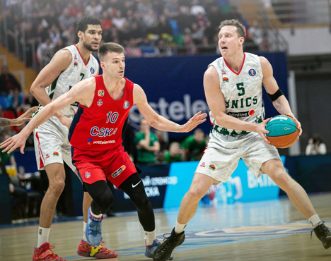 На изображении два баскетболиста на площадке: один в форме красного цвета, другой в бело-зеленой. Игрок с мячом ищет возможность для передачи, другой защищается. На фоне видны зрители и спортивная реклама