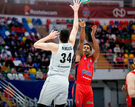 Баскетбольный матч: игрок в красной форме кидает мяч, пытаясь забросить его через игрока в белой форме с номером 34. На заднем плане трибуны с болельщиками