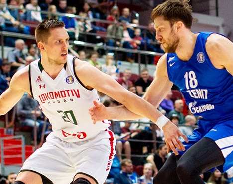 Два баскетболиста в игре: игрок в белой форме Локомотива с мячом против спортсмена в синей форме Зенита. Они находятся на площадке, вокруг наблюдают зрители