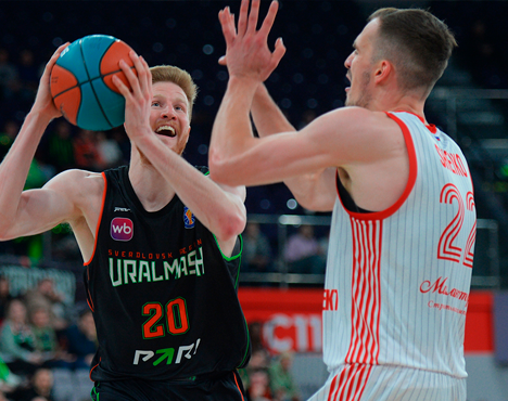 На изображении два баскетболиста в моменте игры. Один в черной форме команды Уралмаш с мячом, готовится сделать бросок. Второй, в белой форме с красными полосами, пытается его остановить. Атмосфера напряженной игры