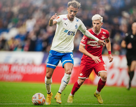 На изображении два футболиста во время матча: игрок в бело-синей форме с мячом и соперник в красной форме. Они находятся на поле, на заднем плане видны трибуны и рекламные баннеры