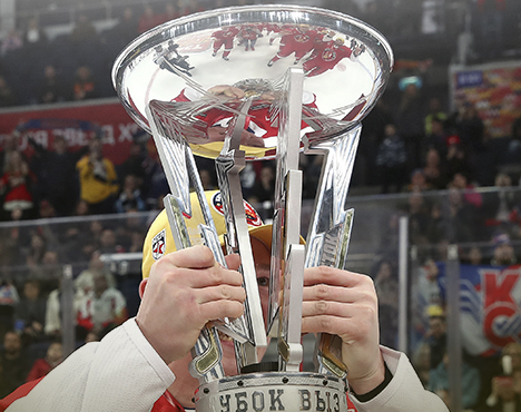 Хоккеист держит блестящий серебристый кубок с выгравированной надписью Кубок Вызова. На заднем плане размыленные зрители на трибунах. Кубок имеет оригинальный дизайн с расходящимися вверх линиями