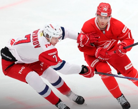 На изображении два хоккеиста соревнуются за шайбу на льду. Один из них в бело-красной форме, другой полностью в красной. Играют на профессиональной арене