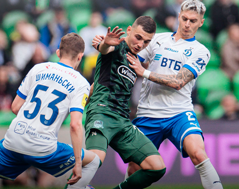 На изображении запечатлен футбольный матч. Игрок в зеленой форме сражается за мяч с двумя игроками в бело-синей форме. Действие происходит на фоне футбольного поля с трибунами