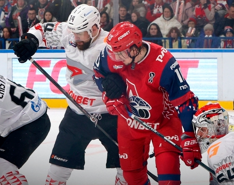 На изображении хоккеисты двух команд в момент борьбы на льду. Игрок в красной форме с клюшкой блокирует соперника, одетого в белую форму. На заднем плане видны болельщики и рекламные баннеры