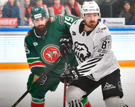 Двое хоккеистов на льду в момент игрового эпизода. Игрок в зелёной форме с эмблемой команды противостоит сопернику в бело-чёрной экипировке. На заднем плане заметны трибуны с болельщиками