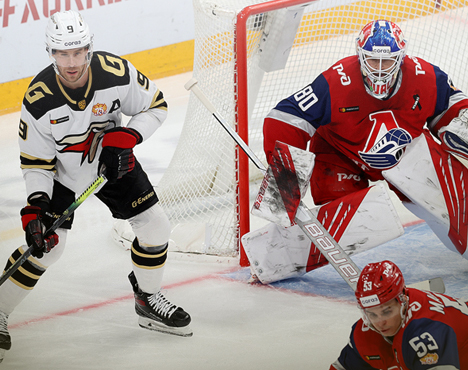 Хоккейный матч: нападающий в белой форме атакует, голкипер в красной форме защищает ворота. Игроки сосредоточены на игре. Динамичный момент у ворот